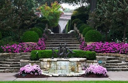 Stone tiered garden with fountain, statues, blooming purple flowers, and steps leading to a building.