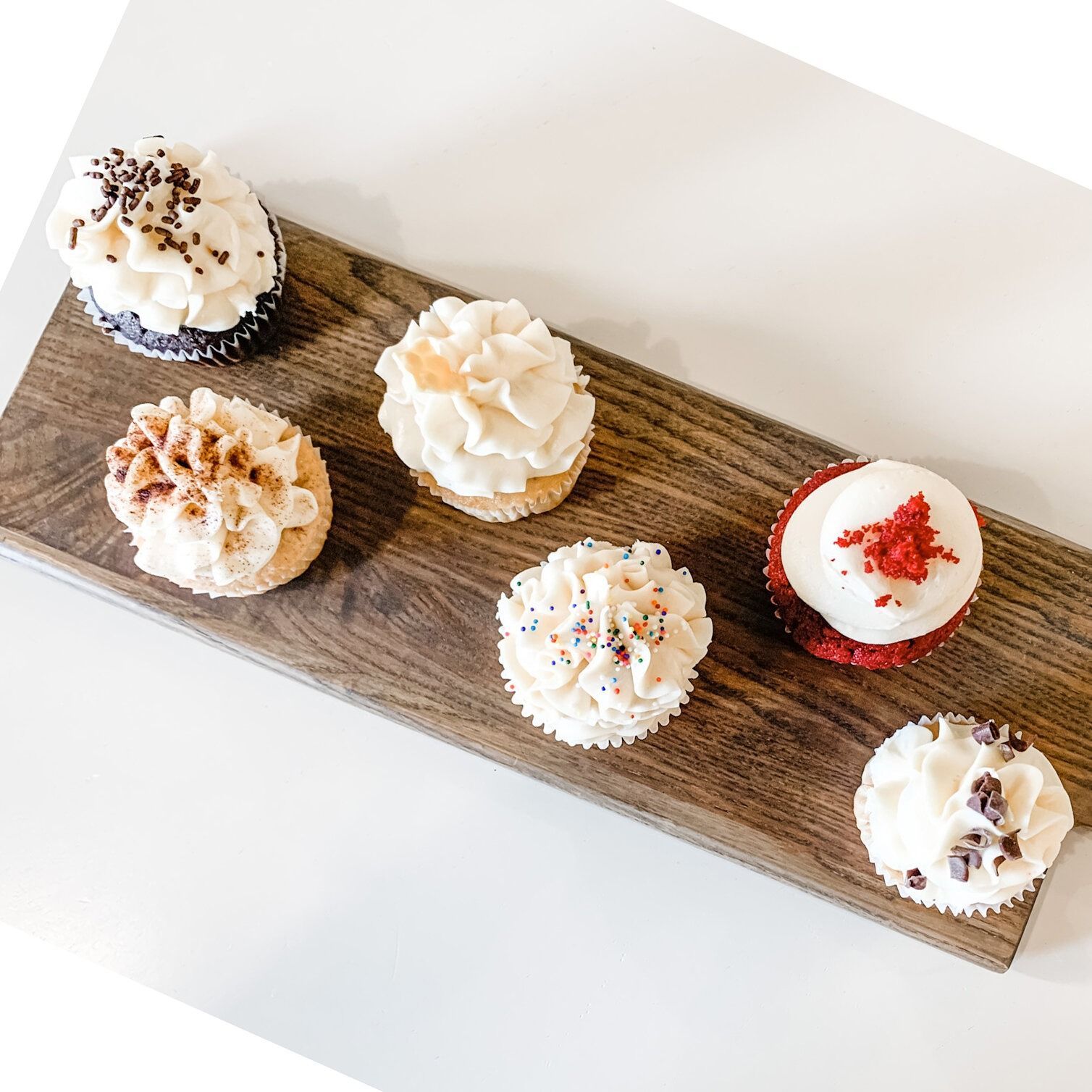 Seven cupcakes with various toppings on a wooden board against a white surface.