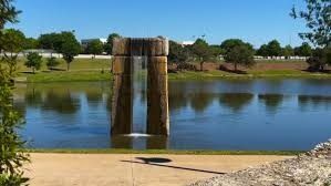 Waterfall sculpture in lake, surrounded by green grass and trees under a clear blue sky.