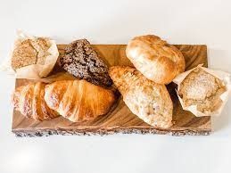 Assortment of baked goods on a wooden serving board: croissant, bread rolls, muffin, and loaf.