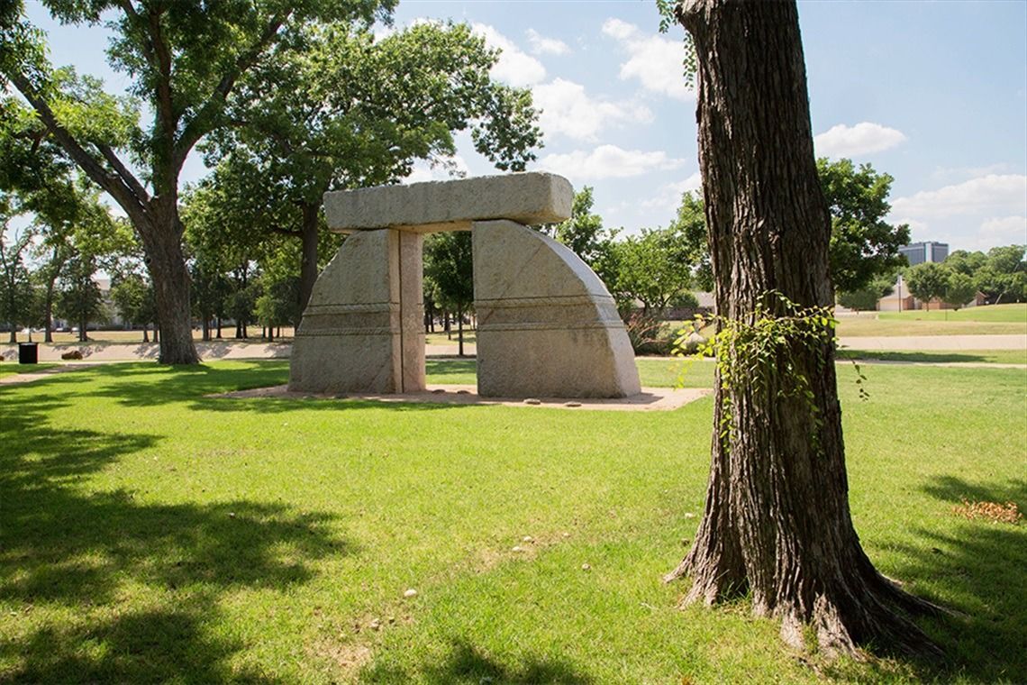 Stone archway sculpture in a park, surrounded by trees and grass, under a blue sky.
