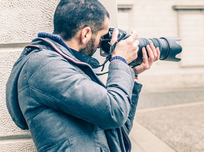 Una persona con una giacca grigia con cappuccio si nasconde dietro un muro mentre scatta una foto con una grande macchina fotografica professionale.