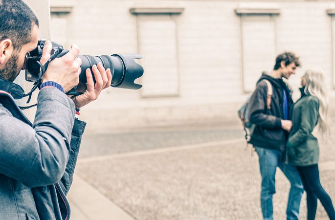 Un fotografo con una giacca grigia usa una macchina fotografica professionale per immortalare una coppia sfocata in piedi in una piazza all'aperto.