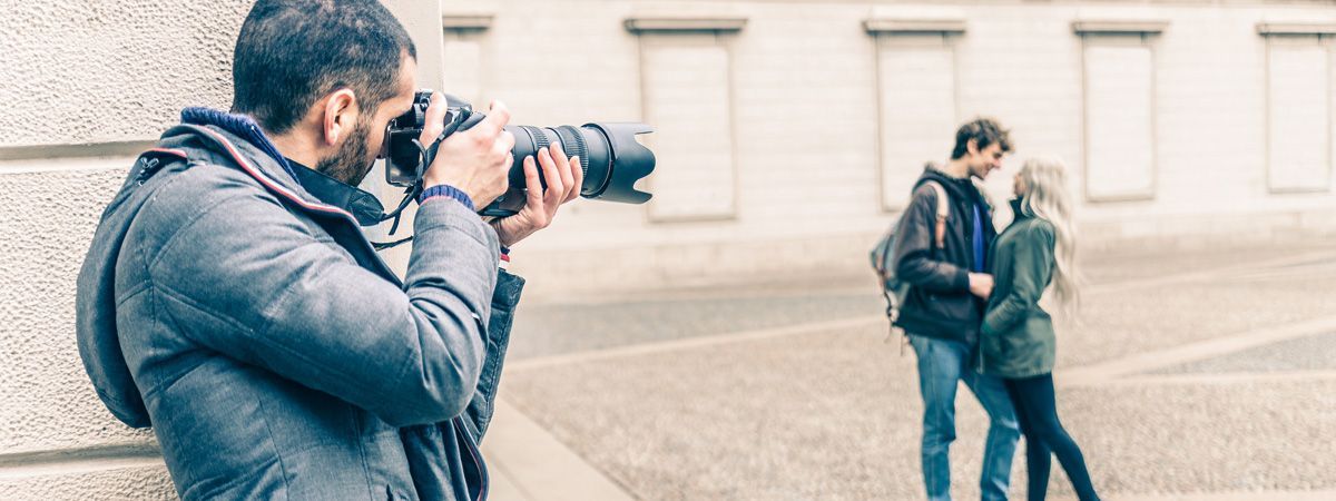 Un fotografo con una giacca grigia è appoggiato a un muro, intento a scattare una foto spontanea a una coppia in piedi all'aperto.