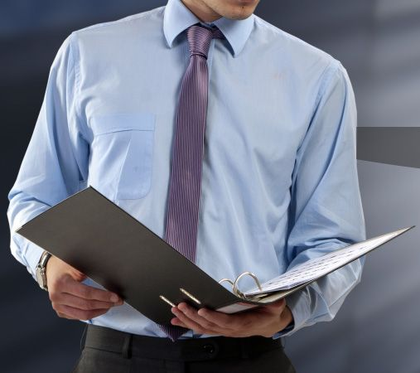 Una persona con una camicia azzurra e una cravatta viola tiene in mano un raccoglitore nero aperto, su uno sfondo neutro.