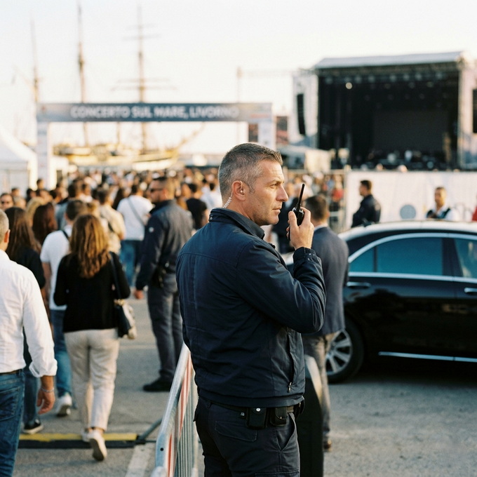 Due persone con indosso camicie blu e bretelle nere sono sedute a delle scrivanie, intente a lavorare su piccoli dispositivi elettronici in un'officina disordinata.