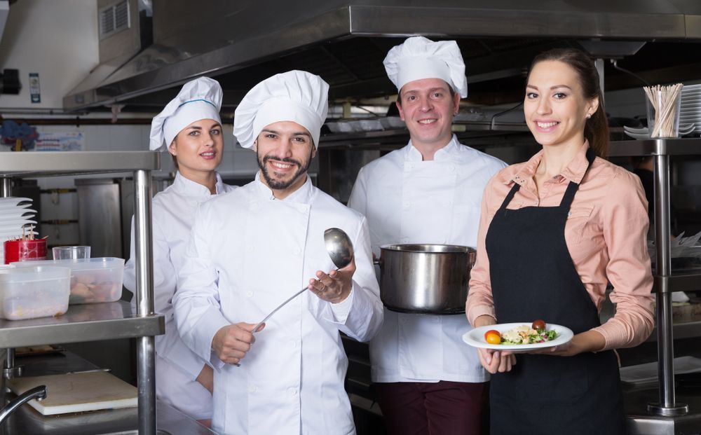 Four restaurant staff pose in a kitchen, chefs holding cooking tools, server holding a plate.