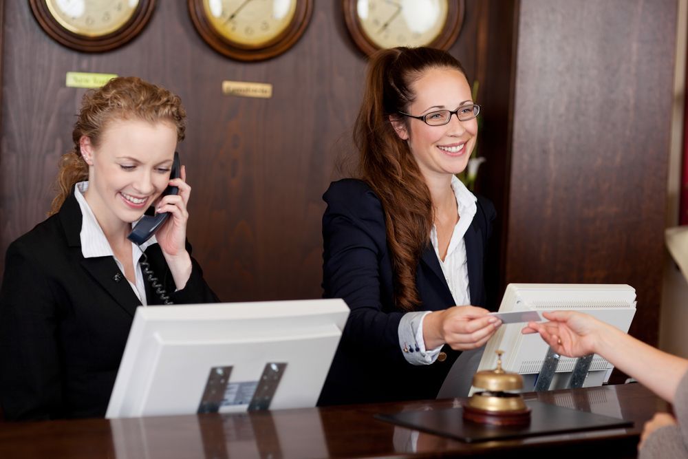 Two hotel receptionists at a desk, one on phone, other handling a card. Wooden desk, world clocks background.
