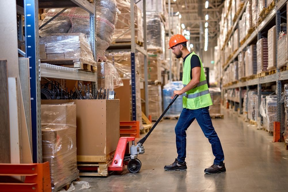 Warehouse worker in safety vest and hard hat operating a pallet jack in a storage aisle.