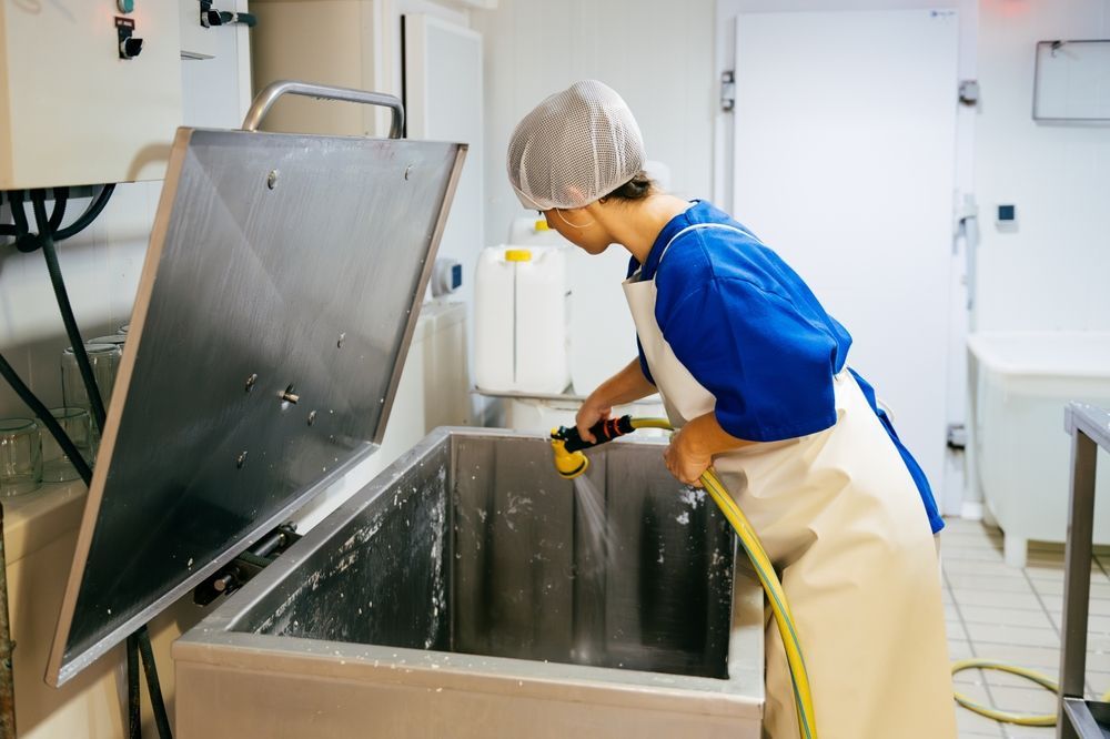 Person cleaning a large, stainless steel vat in a food processing facility, using a yellow hose.