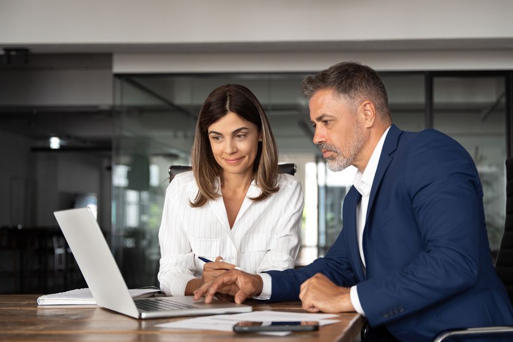 Woman and man looking at a laptop together, both at a wooden table in an office. The woman is pointing at the screen.