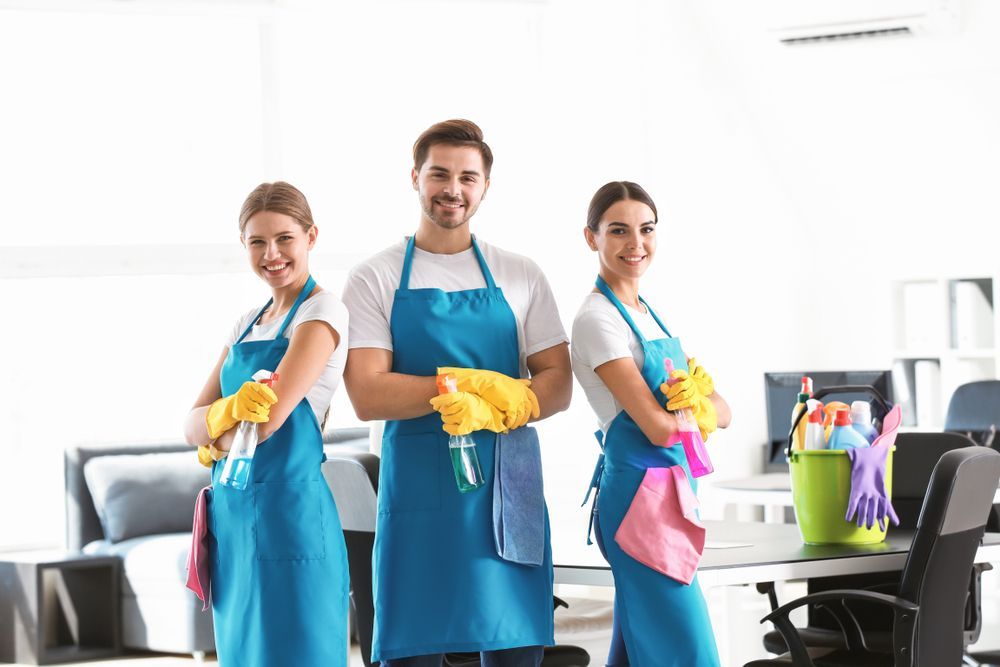 Three cleaning staff in blue aprons and yellow gloves smiling in an office.