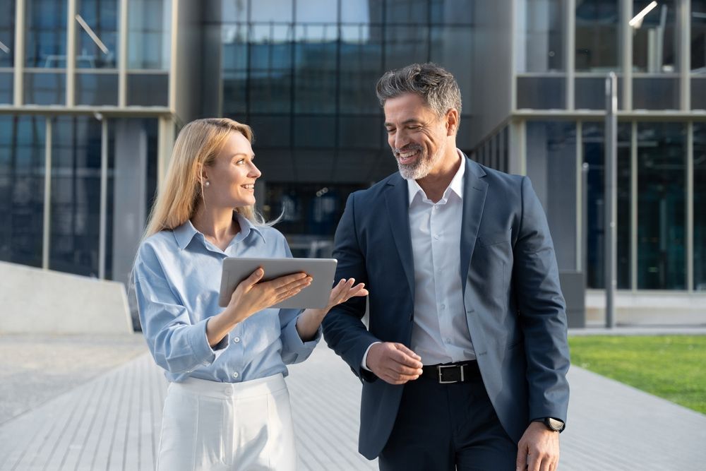 Woman in blue shirt shows tablet to man in suit; they walk outside a modern building, smiling.
