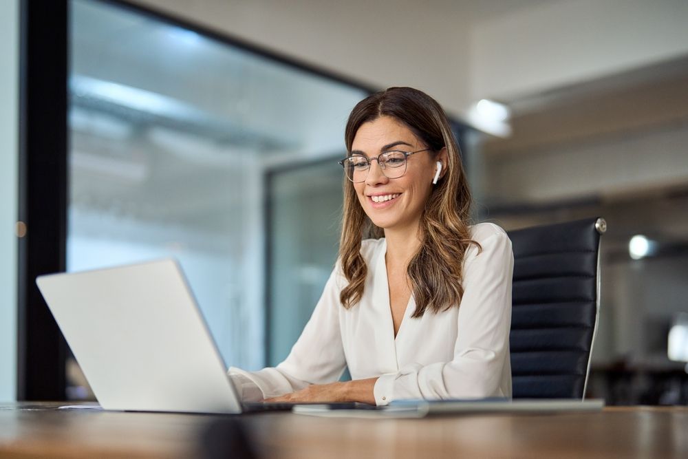 Woman with glasses smiles while using laptop in an office.