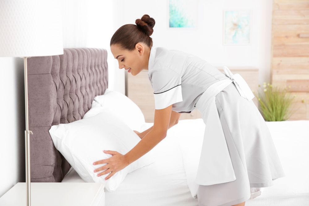 Maid in uniform making a bed, arranging pillows in a light-filled bedroom.