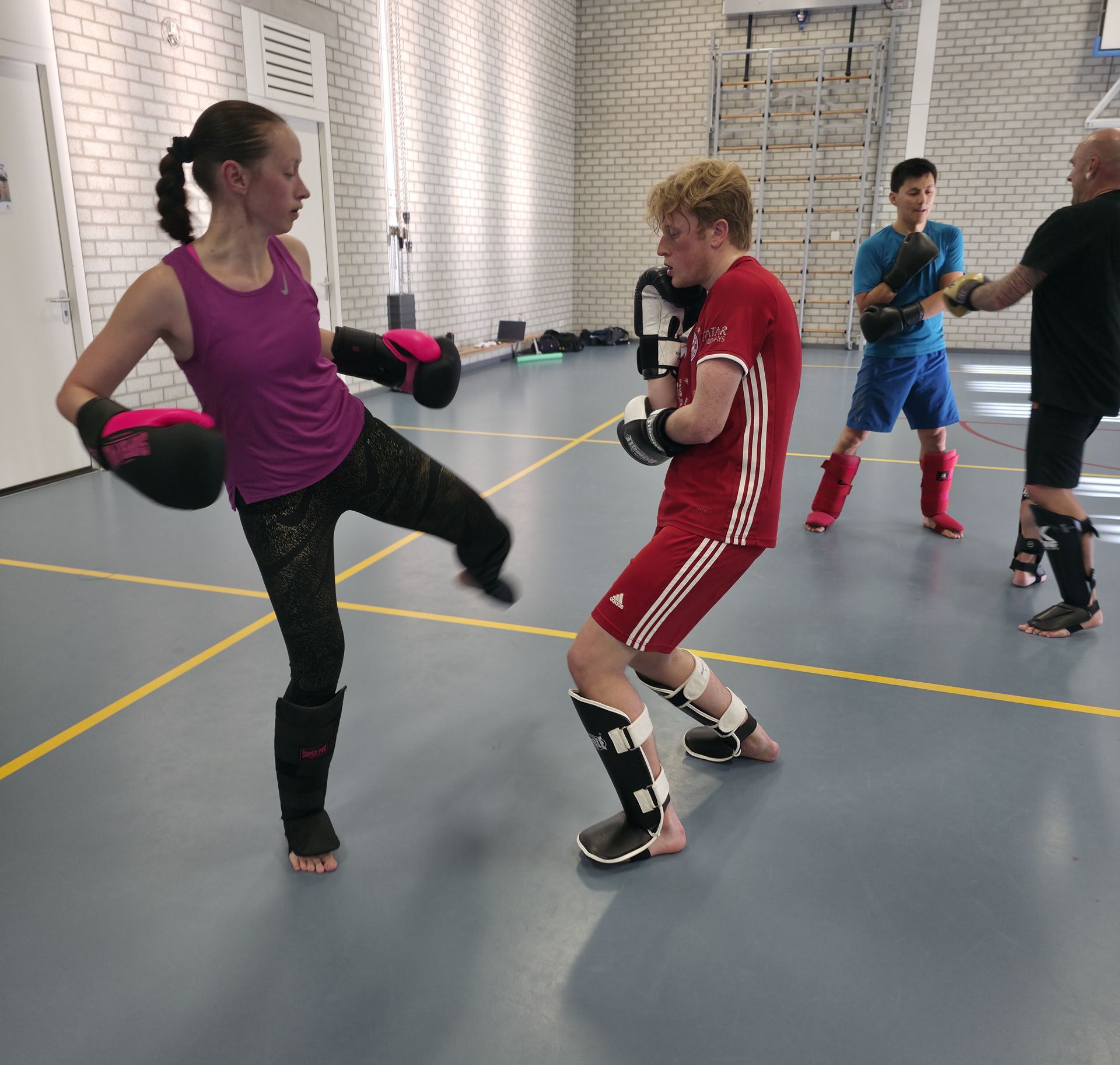 Two people practicing kickboxing in a gym, wearing gloves and shin guards.