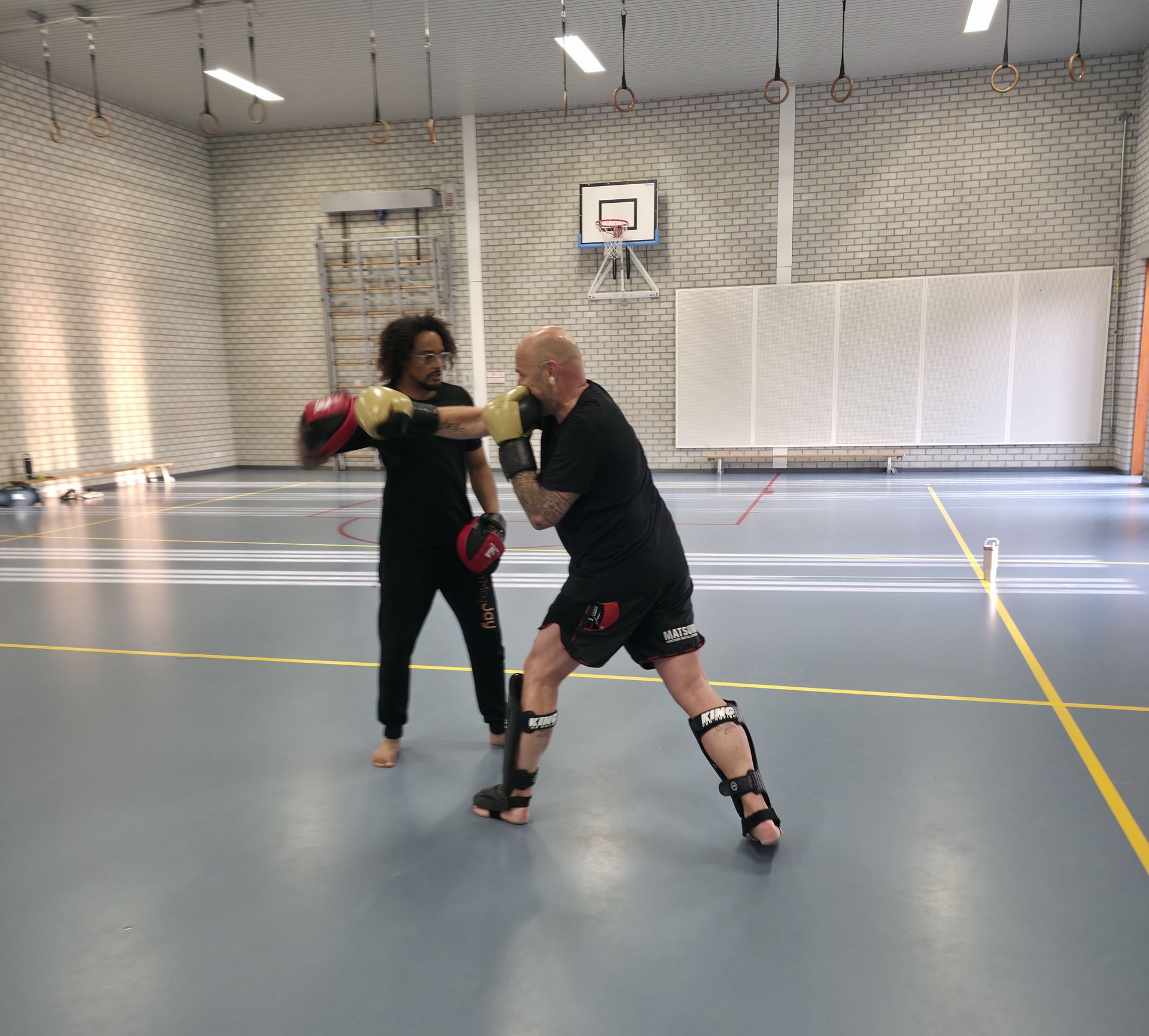 Twee mensen sparren met bokshandschoenen in een binnengymzaal met een basketbalring op de achtergrond.