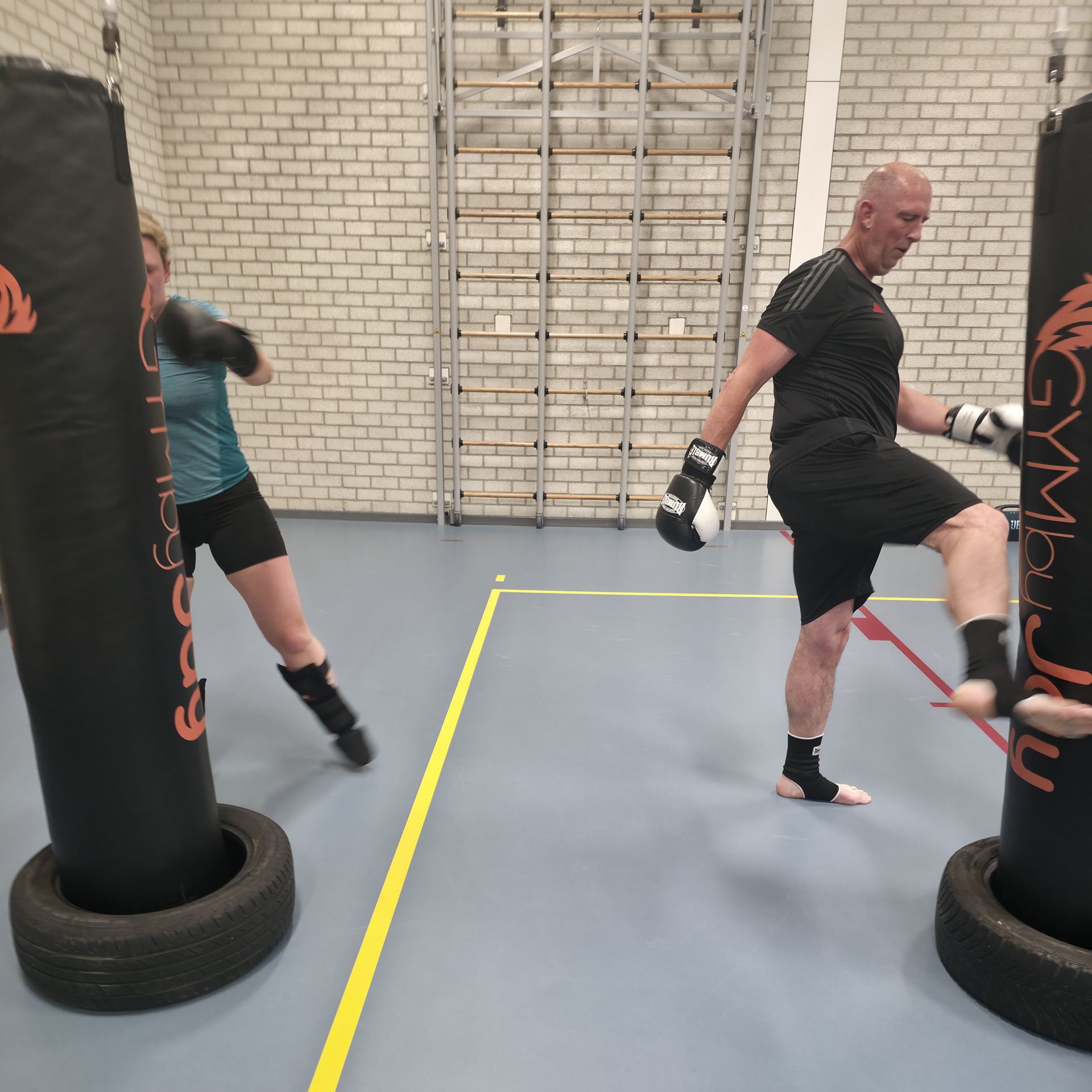 Twee mensen oefenen kickboksen in een sportschool, met handschoenen aan, en trappen in de buurt van zware bokszakken.