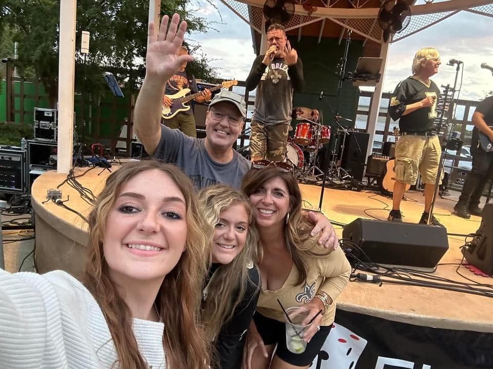A group takes a selfie with a band on stage. People wave, play instruments, and smile. Outdoors.