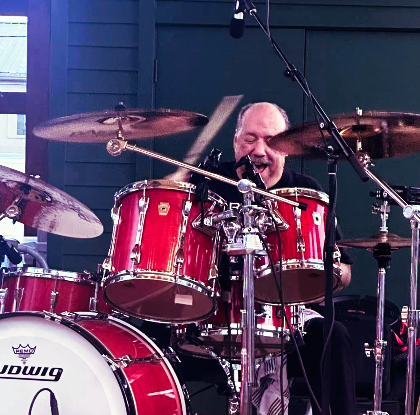 Drummer playing a red drum set outdoors, smiling, with drumsticks raised.