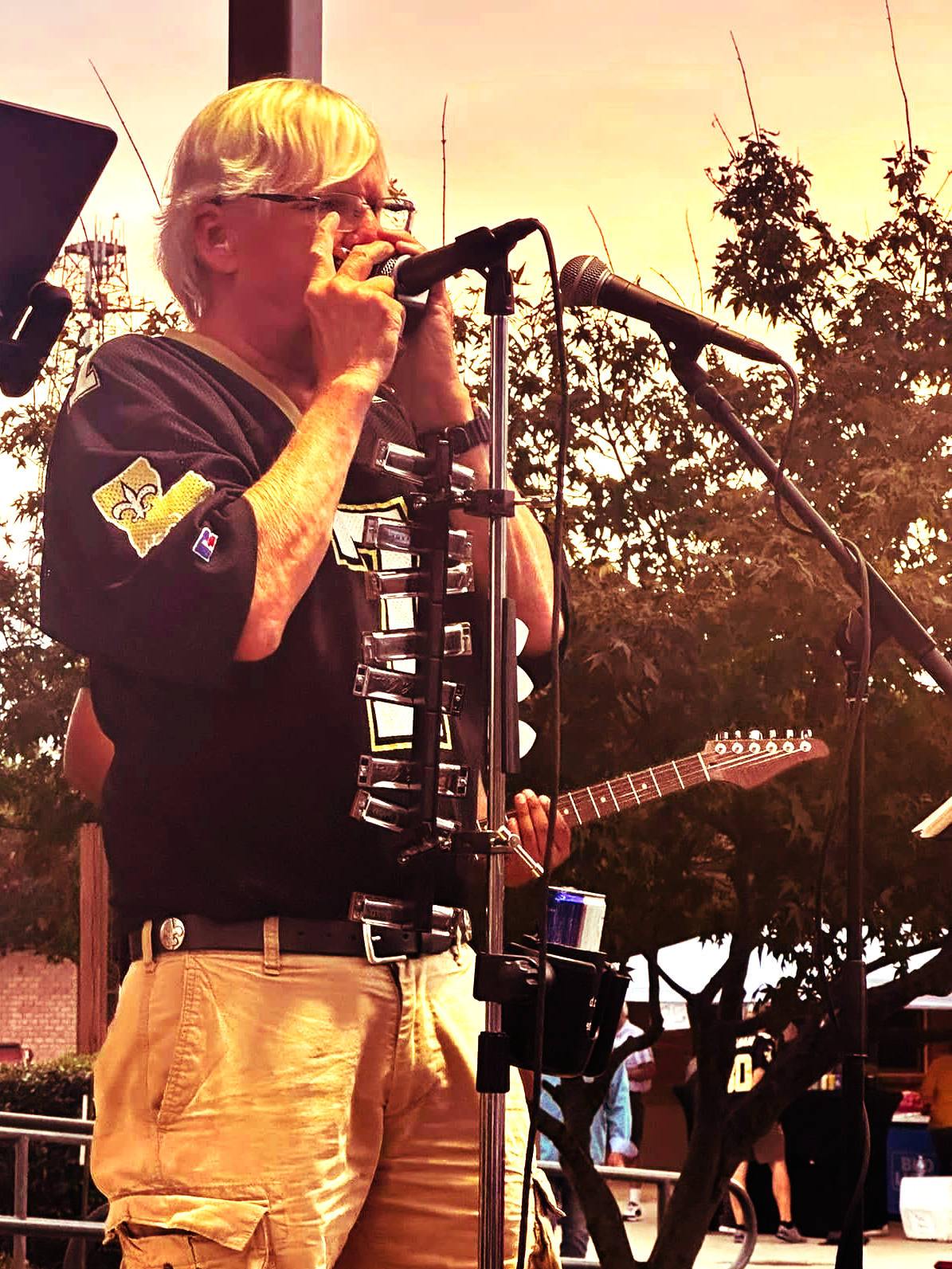 Man playing harmonica and guitar on stage, wearing a Saints jersey, outdoors.