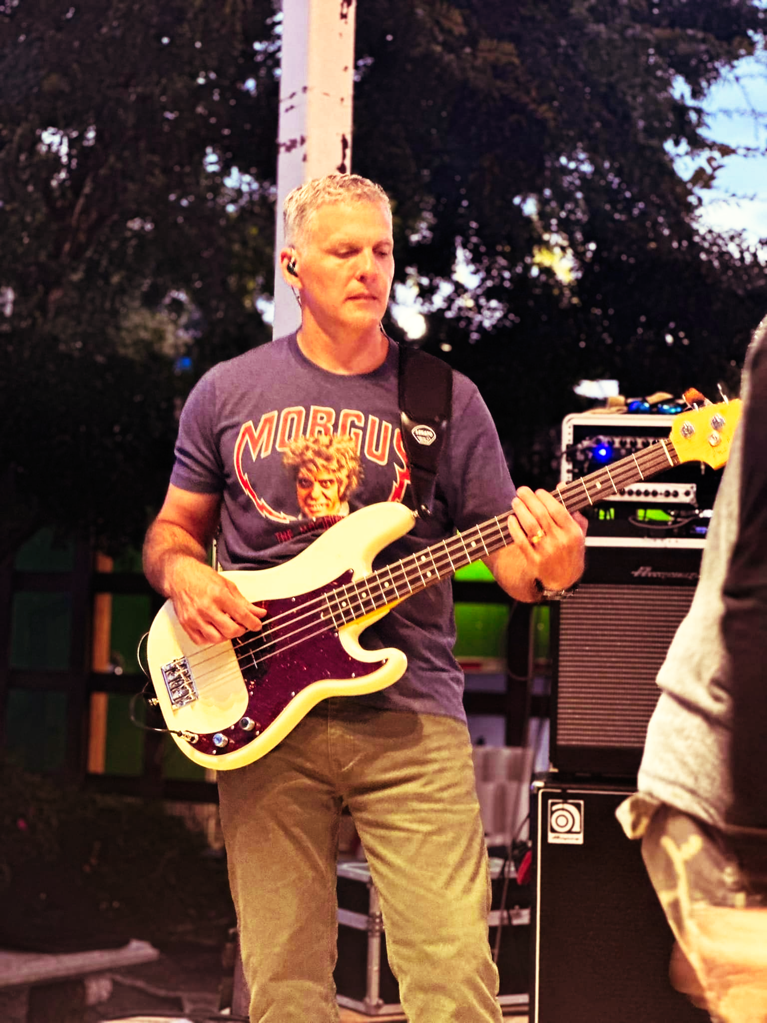 Man playing bass guitar outdoors, wearing a blue t-shirt and green pants.