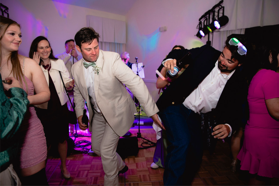 People dancing at a wedding reception. Man in tan suit dances near another in a dark jacket, both smiling. Pink and purple lighting.