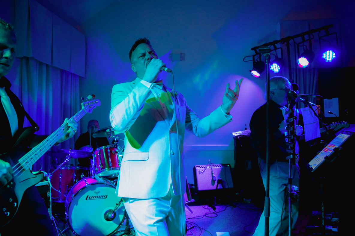 Band performing on stage under blue lights. Singer in white suit holds microphone, gesturing. Other musicians in background.