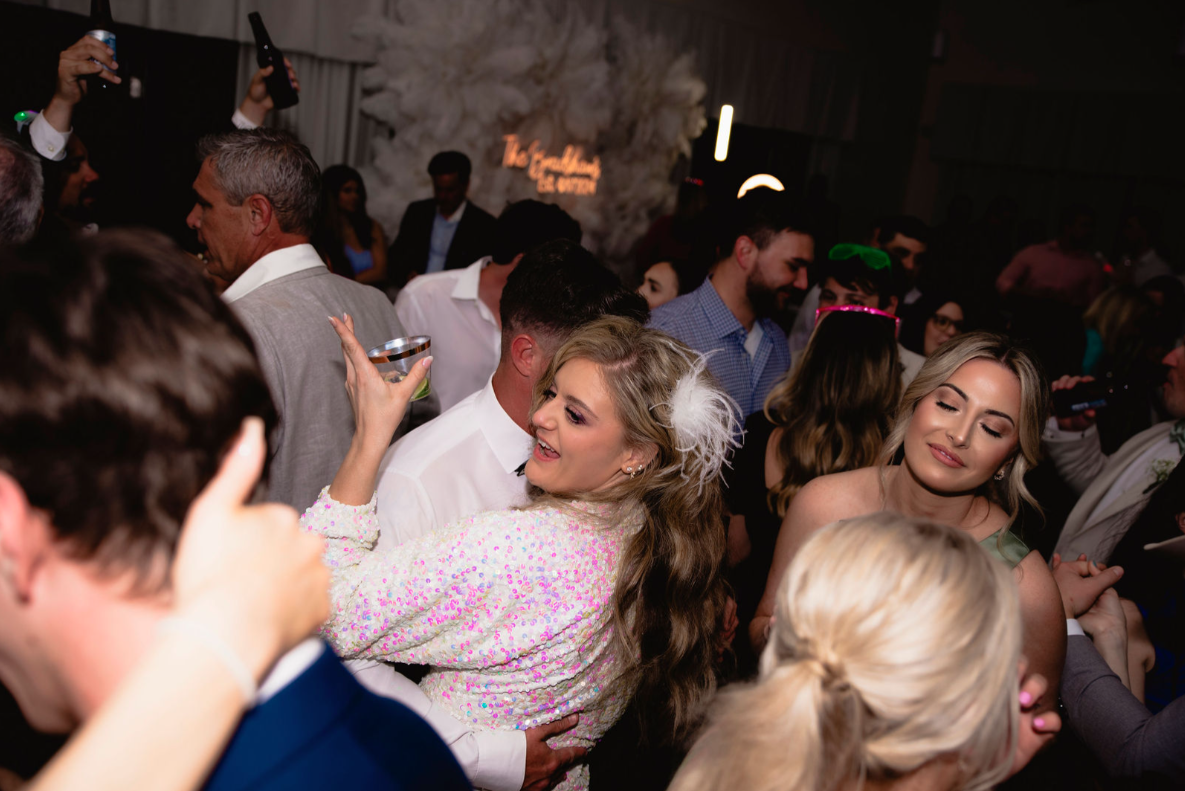 People dancing at a party; woman in sequined jacket smiles, arm around a man. Dark, indoor setting.