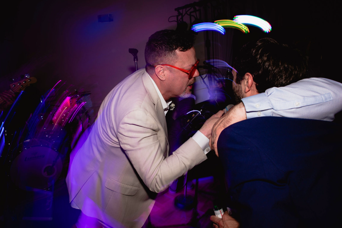 Man in cream suit and red glasses leans in, talking to another man in a blue shirt at a party; dark, with colorful lights.