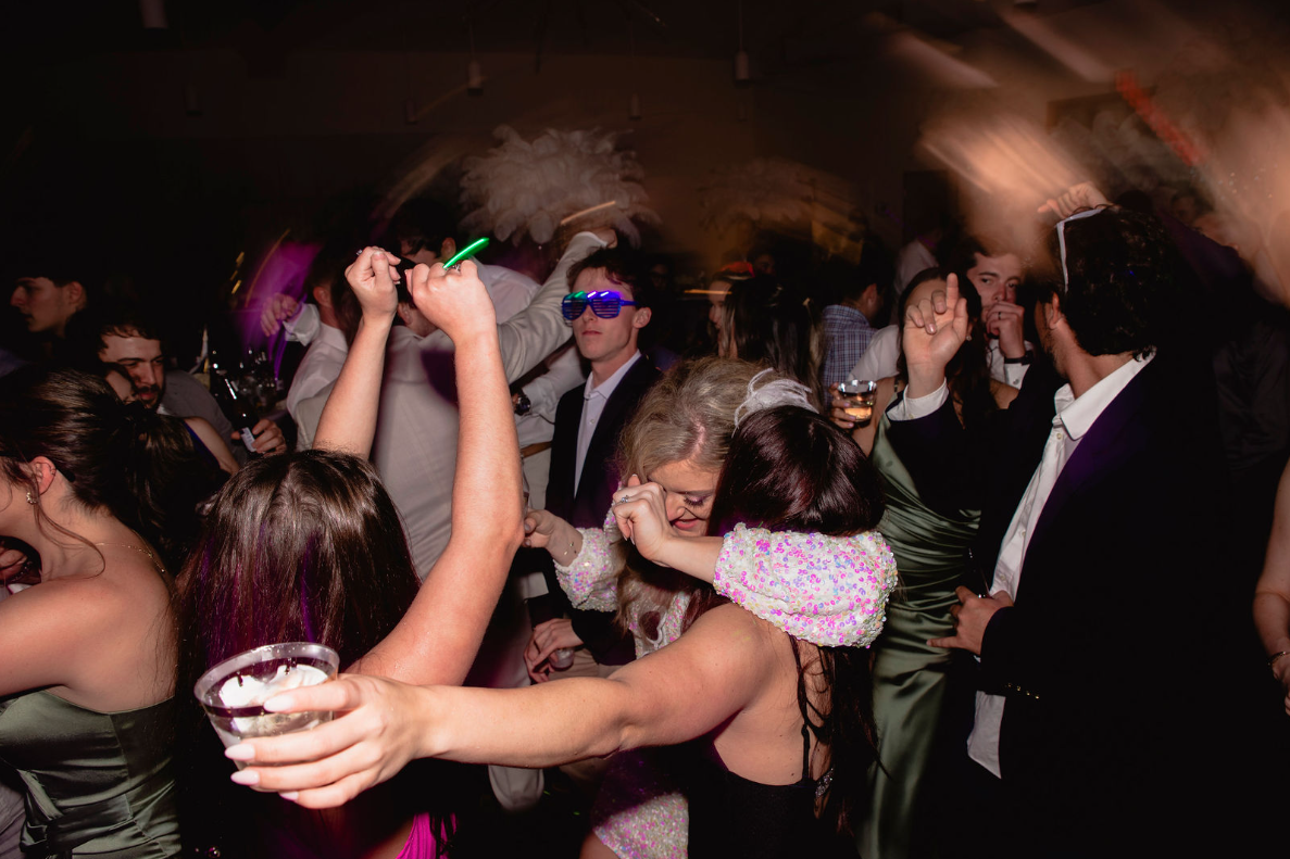People dancing at a crowded, dimly lit party; arms raised, holding drinks, some wearing party accessories.