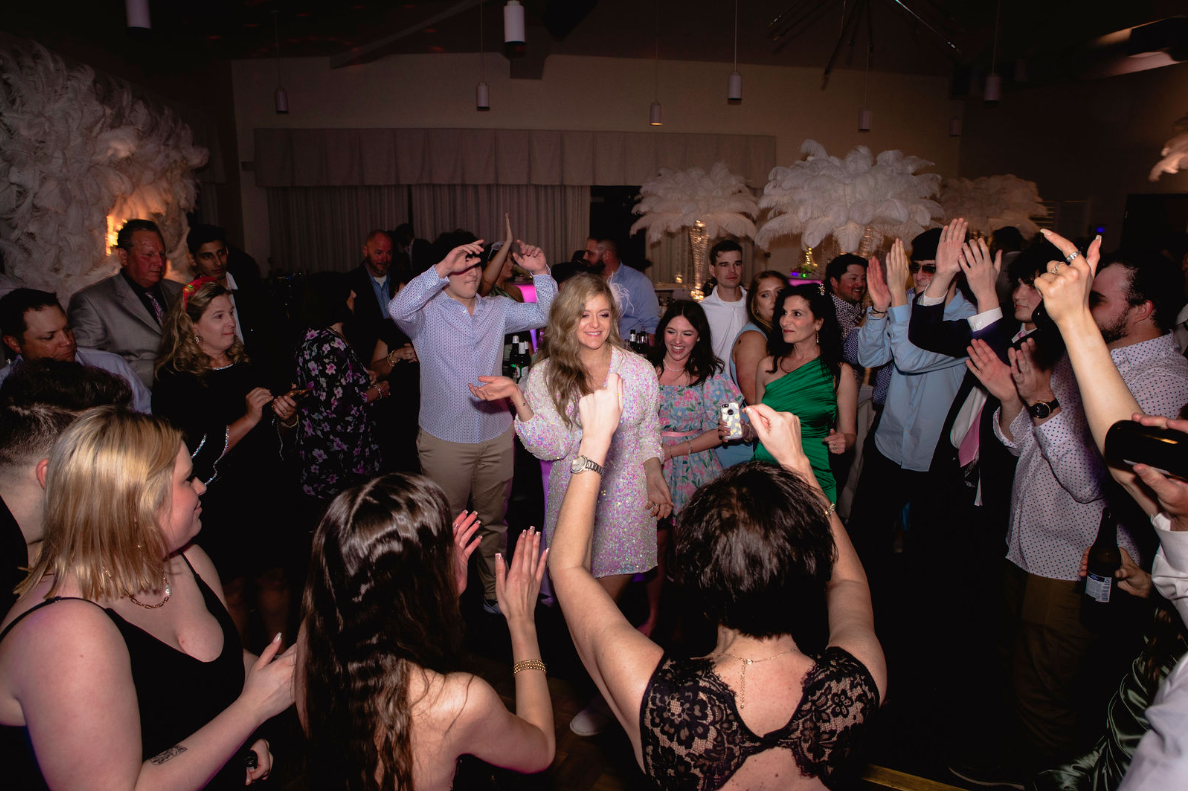 People dancing in a well-lit room. Some have their arms raised. There are white decorations in the background.