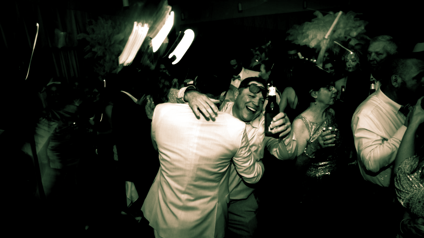 People dancing at a party; one holds a bottle, wearing a mask. Blurred lights, dark setting.