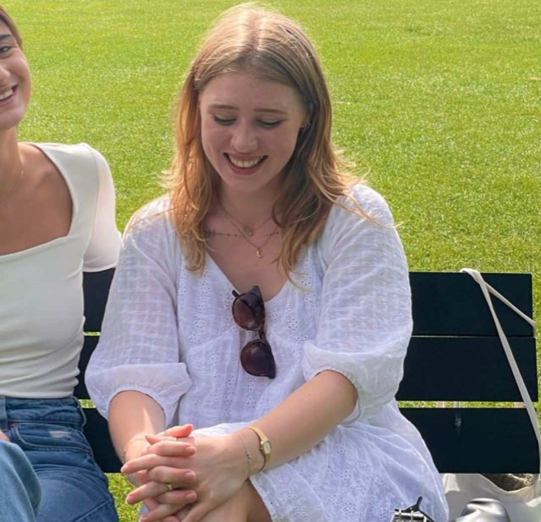 Two women are sitting on a bench with their hands folded and smiling