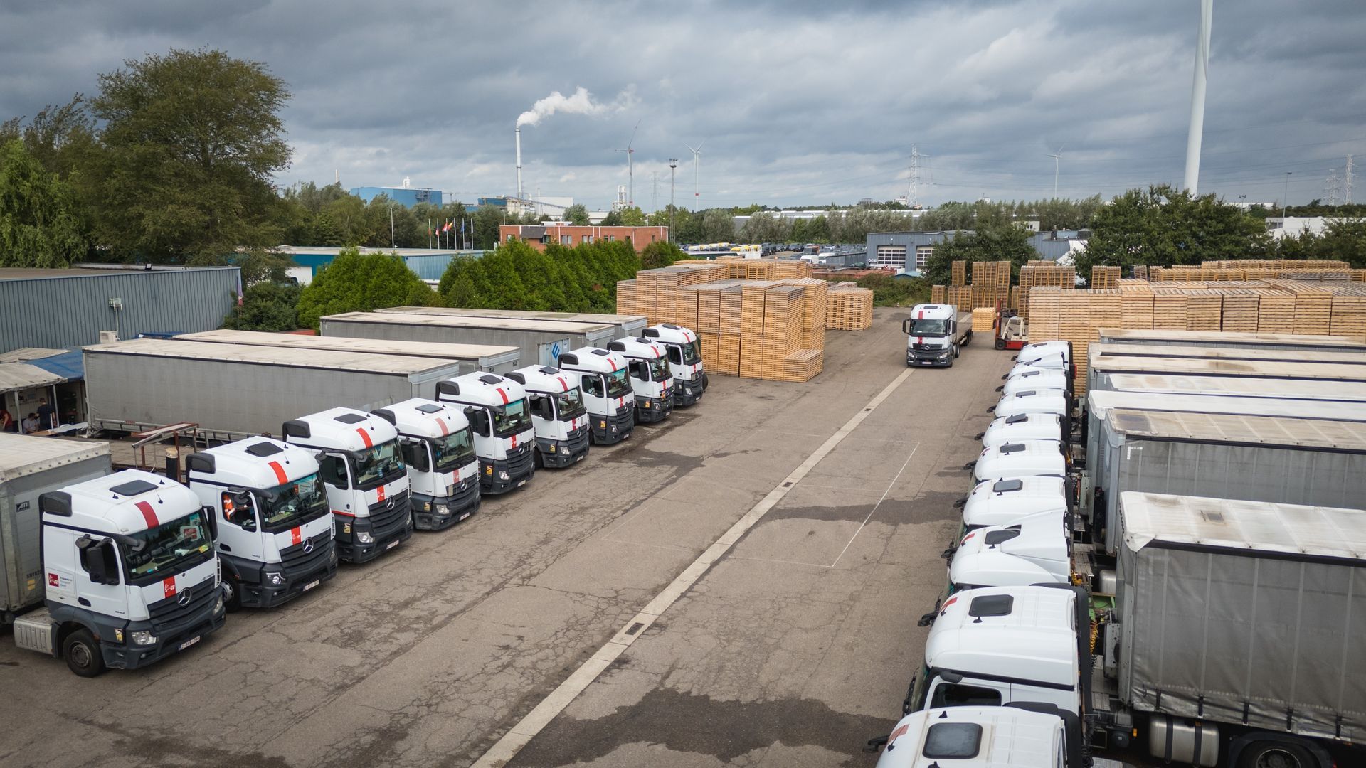 Trucks parked in a loading area; stacks of lumber visible. Overcast sky, industrial setting.