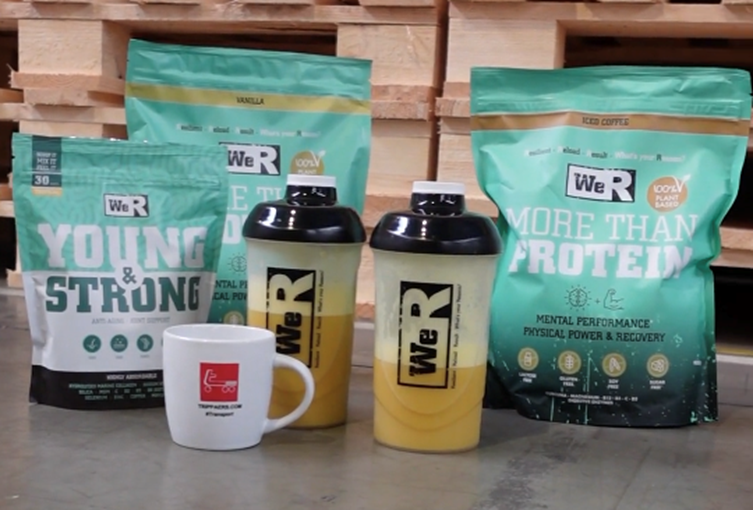 Protein powder bags and shaker bottles on a surface, with a mug, all in front of a wooden wall.