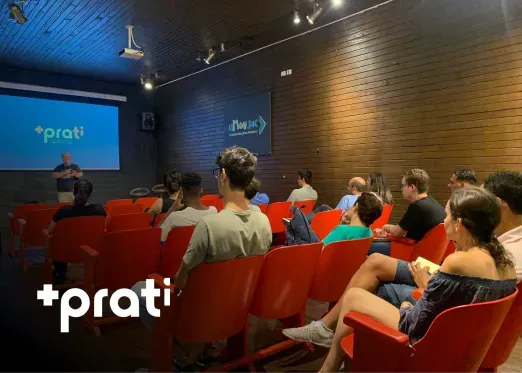 A man gives a presentation in a room with a projector screen. People sit in red chairs, listening. "Prati" logo visible.