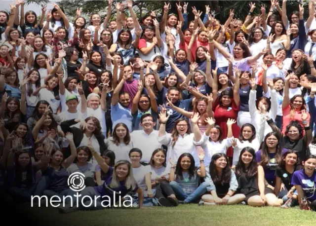 Large group of people with arms raised, outdoors. Mentoralia logo in the corner.