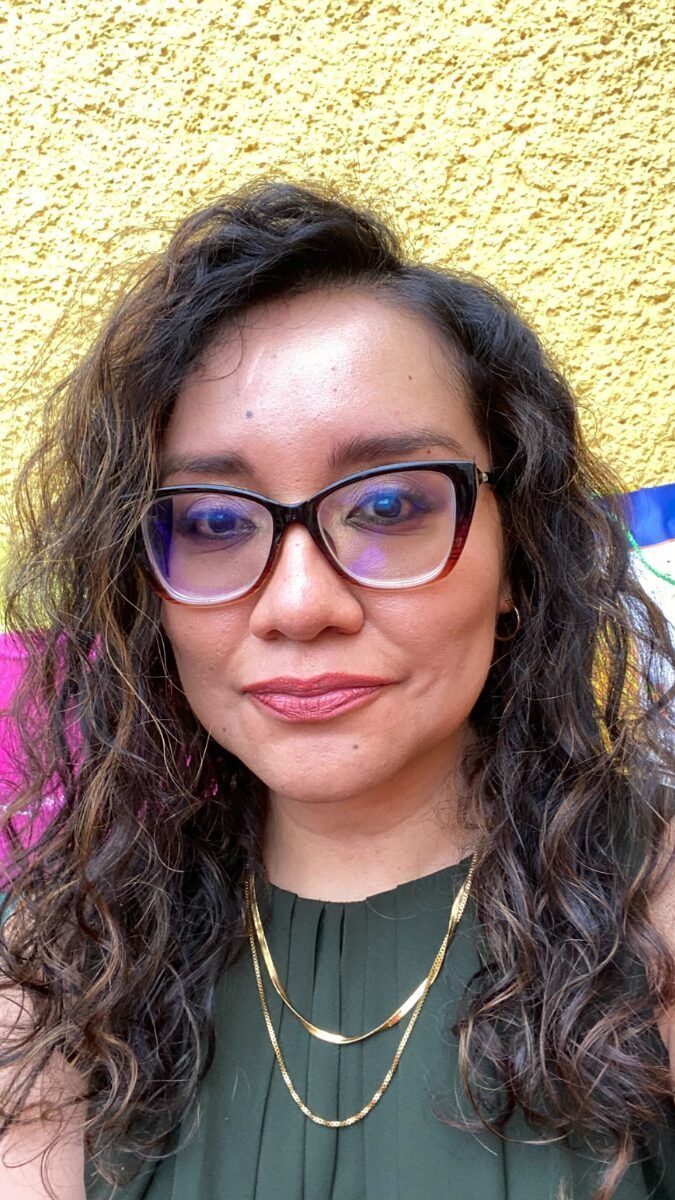 A woman wearing glasses and a necklace is standing in front of a yellow wall.