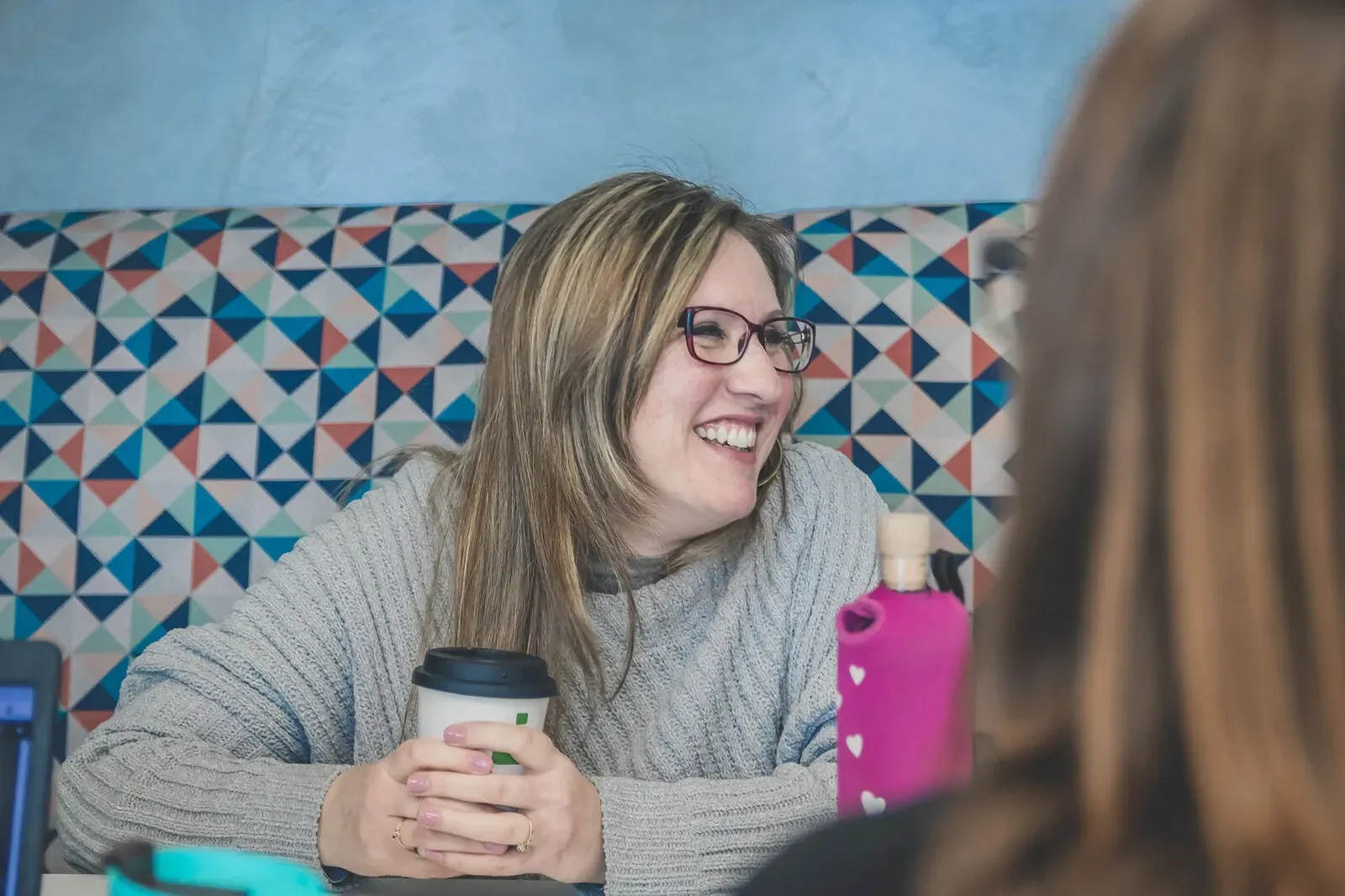 A woman is sitting at a table holding a cup of coffee and smiling.