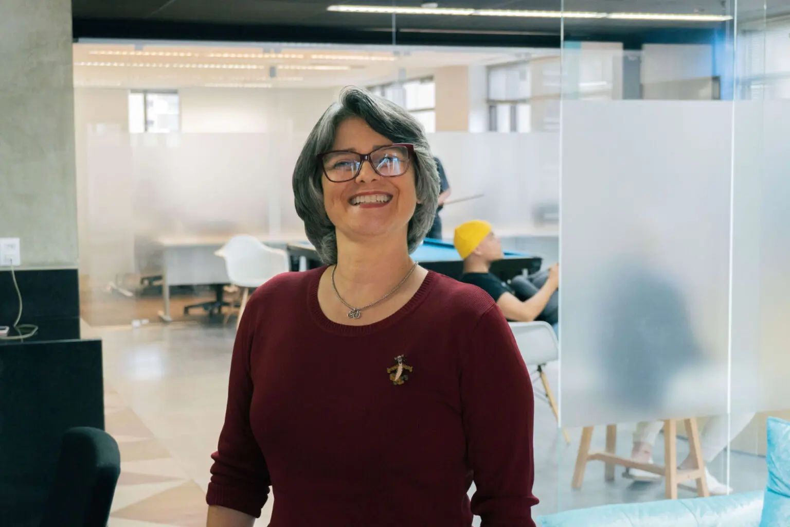 Una mujer con gafas sonríe, vistiendo un suéter rojo, en un espacio de oficina moderno.