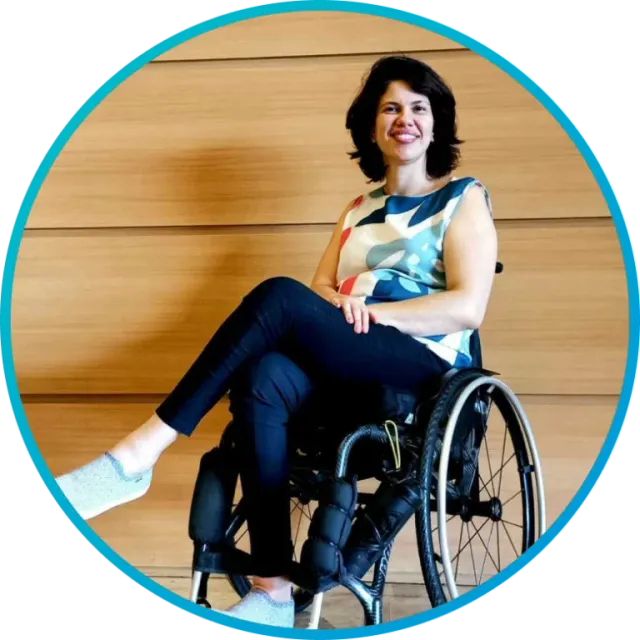 Woman in wheelchair smiles, wearing a patterned top and black pants, in front of a wooden wall.