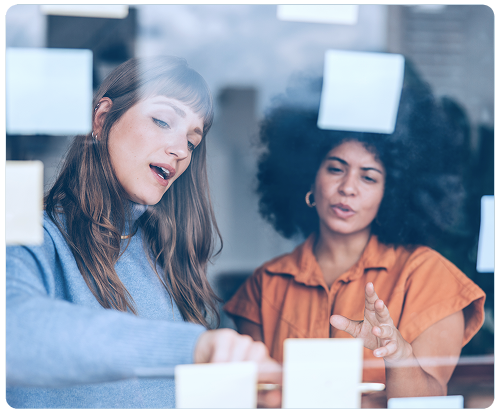 Duas mulheres estão olhando para post-its em uma parede de vidro.