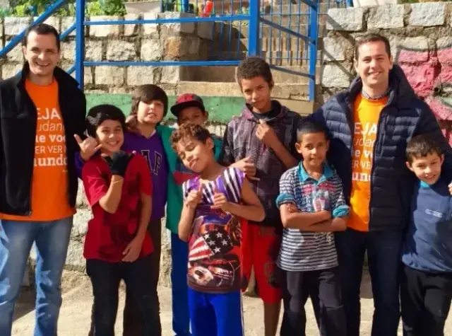 Two men with a group of children, smiling outdoors. They wear matching orange shirts; the setting has a wall and steps.