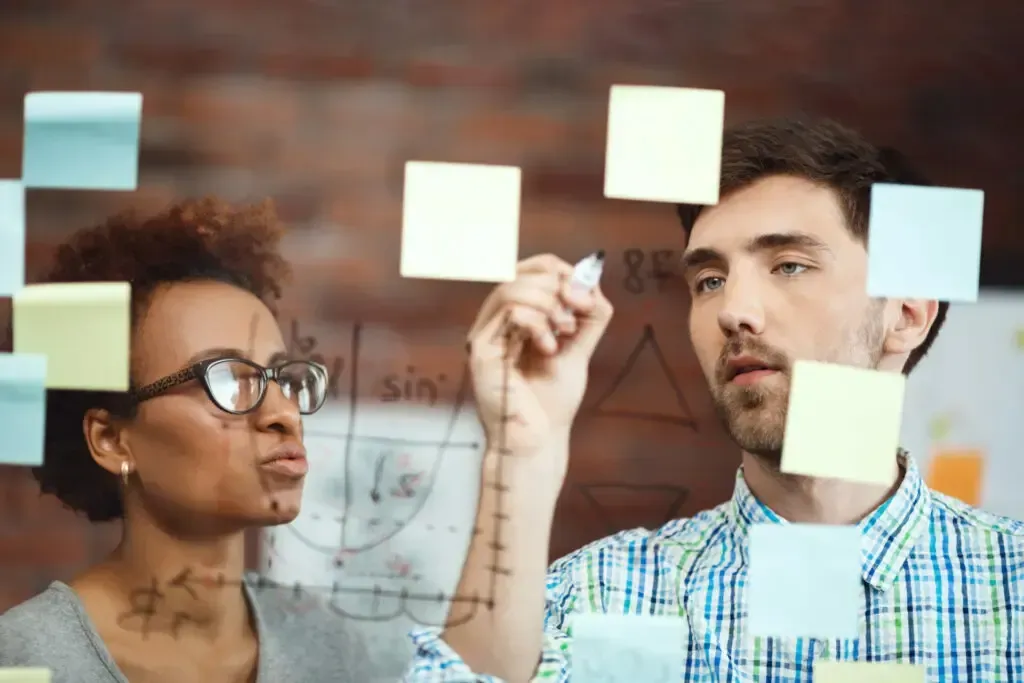 Um homem e uma mulher estão olhando para post-its em uma parede de vidro.