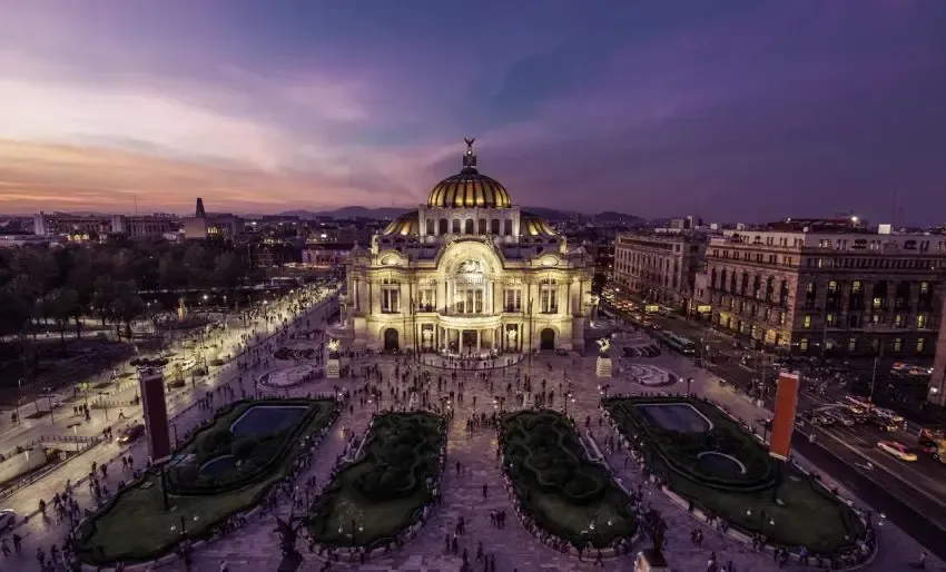 Palacio de Bellas Artes in Mexico City at dusk, illuminated, surrounded by a plaza and buildings.