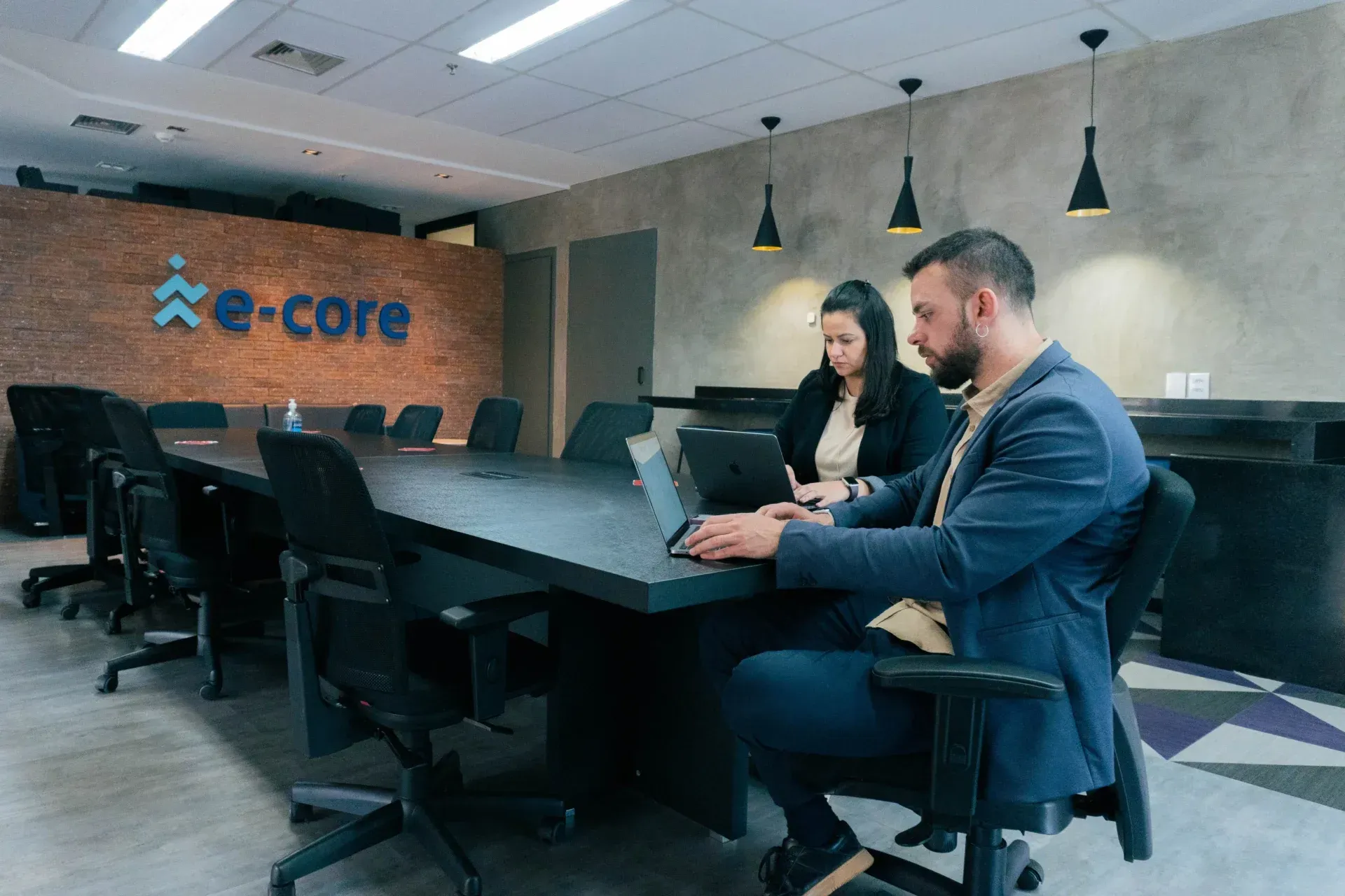 A man and a woman are sitting at a conference table using laptops.
