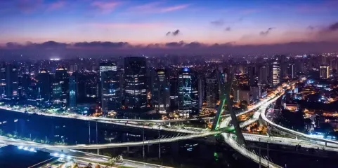 Night view of a city with a lit up bridge and buildings silhouetted against a purple and orange sky.