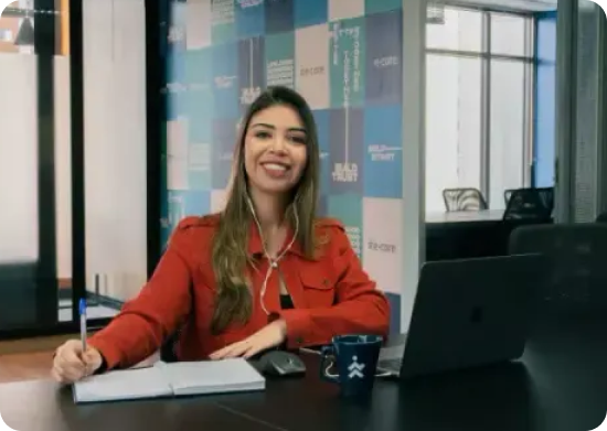 Mulher de camisa vermelha sorrindo em frente a uma mesa, com um laptop, um caderno e uma caneca.