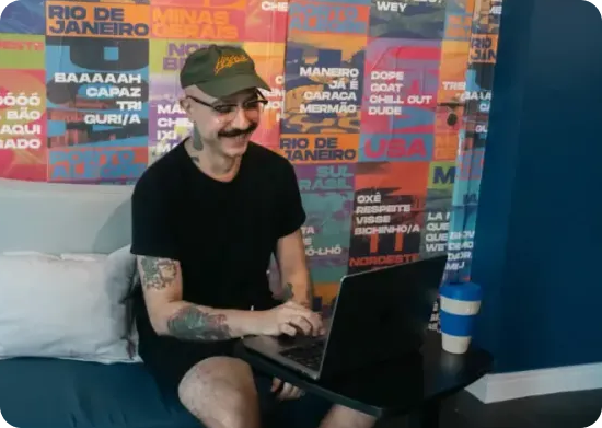 Pessoa com tatuagens, boné e bigode usando um laptop; sorrindo. O fundo apresenta uma arte de parede colorida.